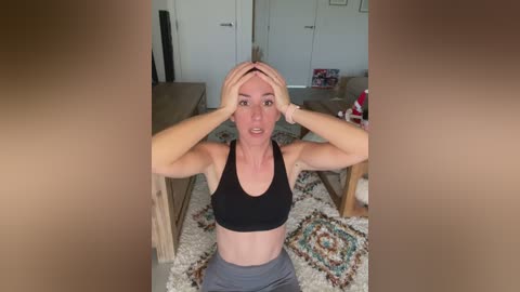 Media: A video of a light-skinned, muscular woman in a black sports bra and gray leggings, sitting on a patterned rug, covering her face with her hands. The background features a wooden table and a white door.