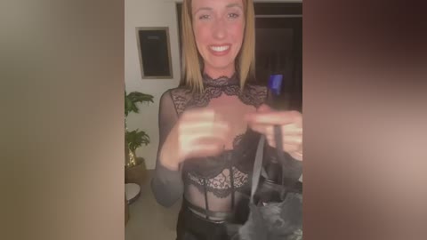 Media: Video of a blonde woman with fair skin, wearing a black lace bralette, smiling, indoors with a potted plant and door in the background.