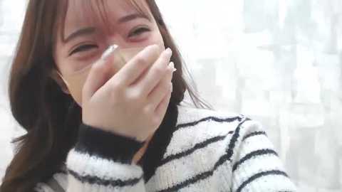 Media: Video of an Asian woman with long, straight brown hair, wearing a striped sweater, covering her mouth with her hand, tears visible, blurred cityscape background.