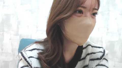 Media: Video of a young East Asian woman with long brown hair, wearing a white and black striped sweater, beige face mask, and light makeup, looking slightly to the right. Background features a blurred, semi-transparent window with a cityscape.