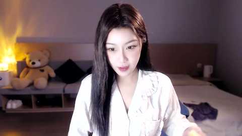 Media: Video of an Asian woman with long black hair, wearing a white button-up shirt, standing in a dimly lit bedroom with a bed, stuffed animal, and lamp in the background.