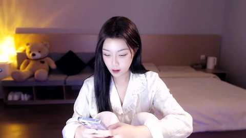 Media: Video of an Asian woman with long black hair in a white robe, looking at a phone, in a dimly lit bedroom with a beige headboard, teddy bear, and nightstand.