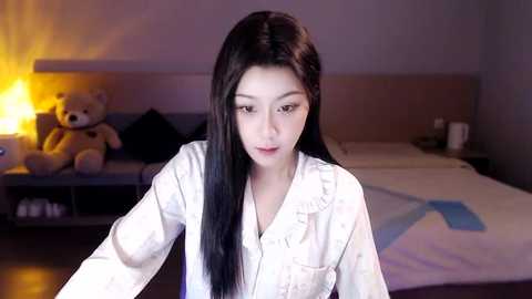 Media: Video of an Asian woman with long black hair in a white nightgown, leaning forward, in a dimly-lit bedroom with a teddy bear, a bed, and a lamp.