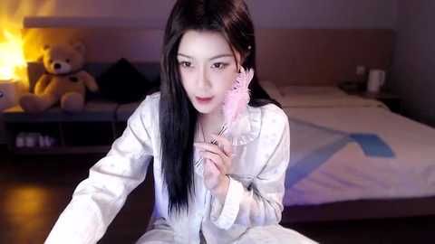 Media: A video of an East Asian woman with long black hair, wearing white pajamas and pink feather earrings, sitting in a dimly lit bedroom with a teddy bear and a bed.