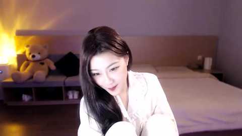 Media: A video of a smiling East Asian woman with long black hair, wearing a white robe, sitting on a bed with a beige headboard. A teddy bear and a lamp are visible in the background.