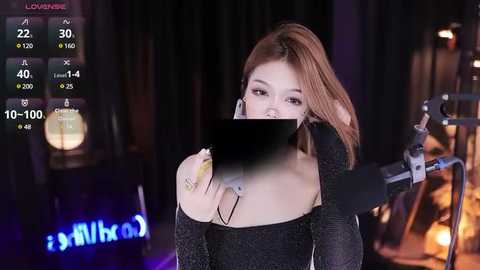 Media: Video of a young Asian woman with light skin and long, straight brown hair, wearing a black off-shoulder dress. She's holding a microphone, her face obscured by a black bar. Background includes a dimly lit room with a candle on a table.