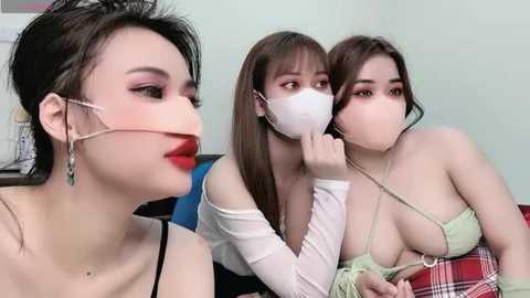 Media: Video of three women in a room; one with red lipstick, two with face masks, wearing revealing outfits, seated in a domestic setting.