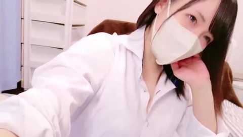 Media: Video of an Asian woman with straight, shoulder-length dark hair, wearing a white long-sleeved shirt, a surgical mask, and a white headband. She rests her chin on her hand, seated in a white room with shelves and a blue curtain in the background.