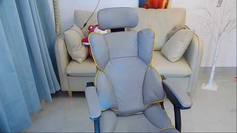 Media: A video of a gray gaming chair with yellow trim in a modern living room with beige couch, blue curtains, and white wall with abstract art.