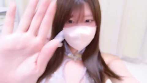 Media: A close-up video of an Asian woman with long brown hair, wearing a white mask, waving her hand to the camera. She's indoors with a soft-focus background.