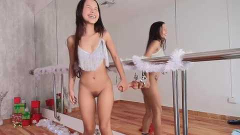 Media: Video of a slender Asian woman with long black hair, wearing a white lace crop top and no pants, smiling and holding hands with a reflection of herself in a mirrored wall.