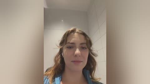 Media: Video of a young Caucasian woman with wavy brown hair, light skin, and a neutral expression. She wears a blue denim shirt, standing in a white-tiled bathroom with a mirror.