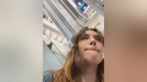 Media: Video of a young woman with long, wavy hair, light skin, and a slight smile, looking upward in a brightly lit room with white walls and shelves.