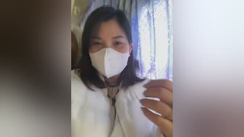 Media: Video of an Asian woman with long black hair, wearing a white surgical mask, white coat, and beige top, holding a syringe, indoors with blurred curtains in the background.