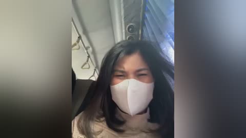 Media: A video captures a woman with long black hair, wearing a white mask, seated in a bus. The background features blurred, indistinct objects.