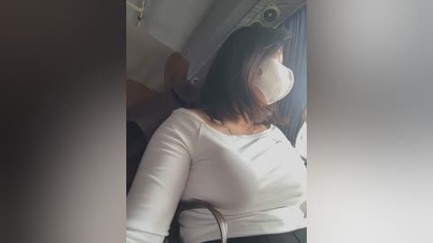 Media: Video of a woman wearing a white face mask and white long-sleeve top, seated in a dark car, with blurred background showing a passenger seat and window.