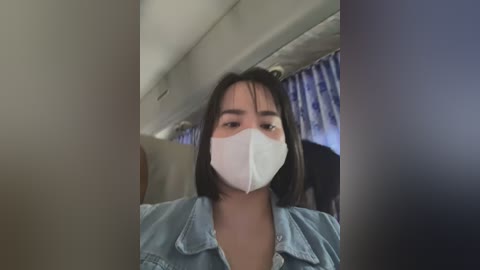 Media: Video of a woman with medium skin tone, shoulder-length black hair, wearing a light blue denim jacket and a white face mask, seated on an airplane, with a blurred cabin background.