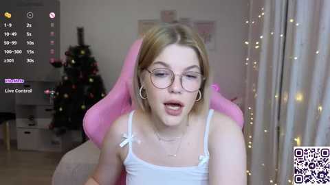 A video of a young woman with fair skin, blonde hair, and round glasses, wearing a white tank top, sitting in a pink gaming chair. A festive Christmas tree and QR code are visible in the background.