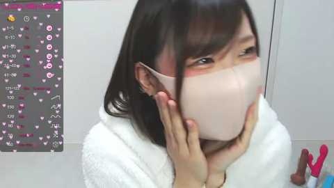 Media: Video of an Asian woman with short black hair wearing a white mask, touching her face, in a white bathrobe. Background shows a white wall with a pink and white heart-patterned smartphone screen.