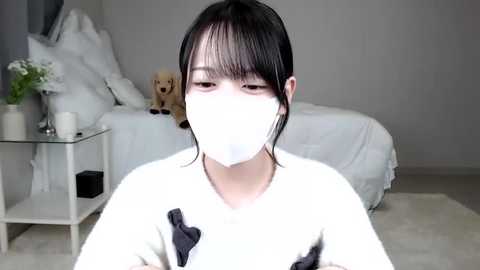 Media: Video of a young Asian woman with short black hair, wearing a white face mask, white sweater, and light-colored pants, sitting in a minimalist, white bedroom with a bed, teddy bear, and glass table.