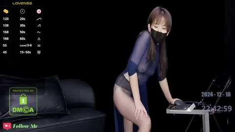 Media: A video of an Asian woman with long brown hair, wearing a sheer blue robe and black face mask, bending over a desk in a dimly-lit room, with a black couch and a green logo in the background.