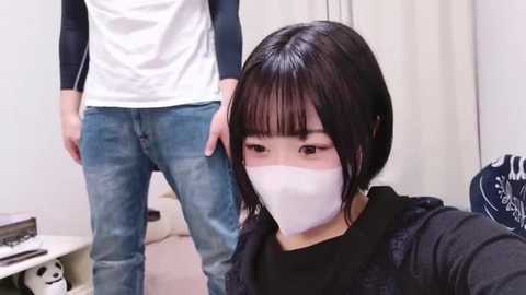 Media: Video of a young East Asian woman with short black hair, wearing a white mask, black top, and black jacket, taking a selfie. Background shows a man in a white shirt and blue jeans.