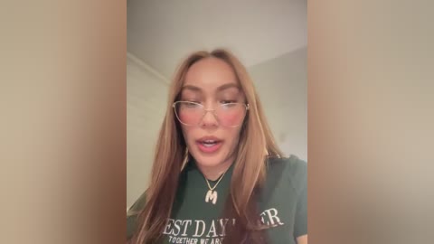 Media: A video of a young woman with long, straight, light brown hair, wearing glasses and a dark green T-shirt with \"BEST DAY EVER\" text. She has a neutral expression, and the background is a blurred indoor space.