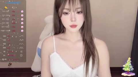 Media: Video of a young Asian woman with long brown hair, fair skin, wearing a white lace camisole, sitting indoors with a plush teddy bear behind her.