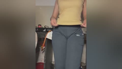 Media: Video of a person lifting a yellow tank top, revealing tight, gray athletic pants. Background includes a countertop with kitchen items and a red canister.