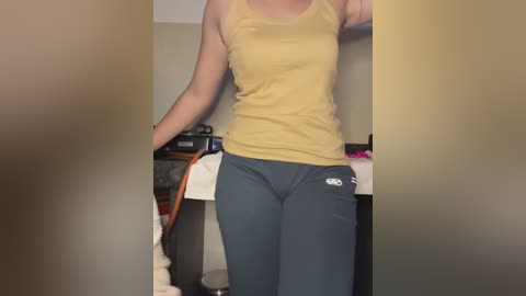 Media: Video of a woman in a mustard-yellow tank top and dark blue sweatpants, standing in a cluttered room with a television and a small white cat.