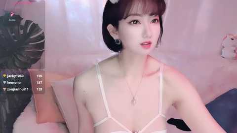 A video of a young Asian woman with fair skin, black bob hair, and pink lipstick, wearing a white bra, sitting on a bed with a plush toy and digital overlays.