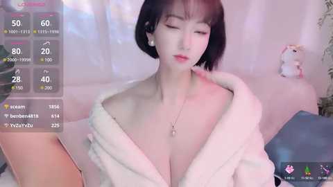 Video of a young Asian woman with a short bob haircut, wearing a white bathrobe, sitting on a bed with a plush toy. Soft pink lighting.