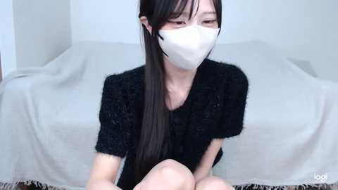 Media: Video of an East Asian woman with long black hair, wearing a white face mask, black knit sweater, and sitting on a white couch.