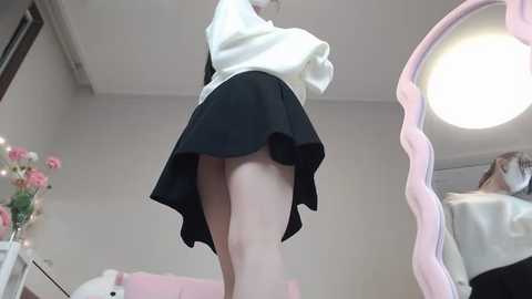 Media: Video of a light-skinned person in a black skirt lifted, revealing bare thighs, standing in a pink-framed mirror, surrounded by flowers and a bed in a brightly lit room.