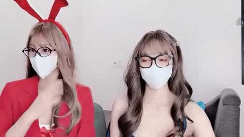 Media: Video of two Asian women with long hair, wearing white masks, black-framed glasses, and red bunny ears, in red and white outfits, sitting on a gray couch.