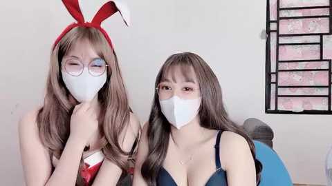 Media: Video of two Asian women in bunny costumes, wearing white face masks, sitting indoors. One has red bunny ears and glasses; the other has long brown hair.