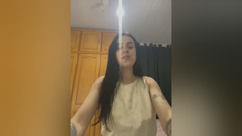 Media: A video of a young woman with long dark hair, wearing a sleeveless beige top, standing in a dimly lit room with wooden cabinets and black curtains.