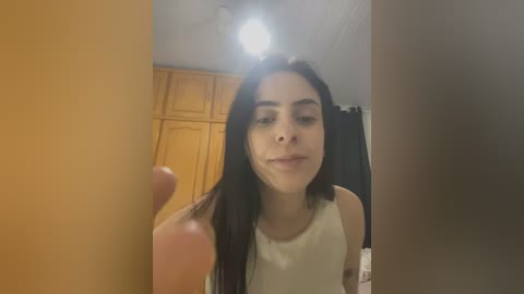 Media: A video of a young Latina woman with long black hair, wearing a white tank top, taking a selfie in a dimly lit room with wooden cabinets and a black curtain in the background.