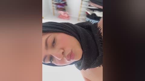 Media: A video of a woman with fair skin and a black hijab, wearing subtle makeup, standing in a room with clothes hung on a rack in the background.