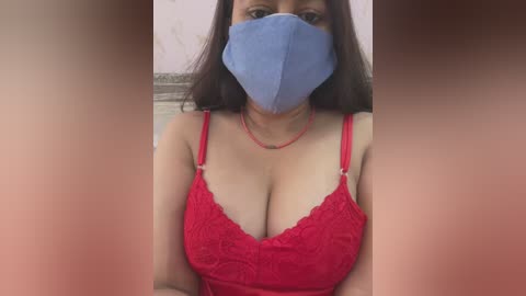 Media: Video of a woman with long dark hair, wearing a blue face mask, a red lace bra, and a red necklace, indoors with blurred background.