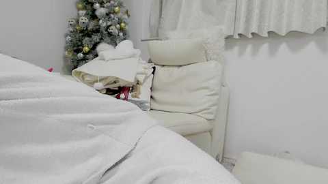 Media: Video of a minimalist, white-walled bedroom with a snow-covered Christmas tree and a pile of white pillows and blankets.