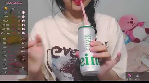 Media: A video of an East Asian woman with long black hair, wearing a white t-shirt, drinking a can of \"Eliker\" energy drink. Background features a plush pink bear and a plush dog.