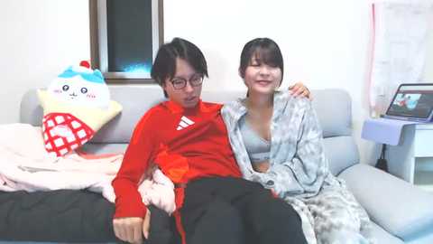 A video of two East Asian women, one in a red jacket, the other in a patterned robe, sitting on a light gray couch, surrounded by plush toys, including a Pikachu pillow.