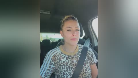 Media: Video of a blonde woman with light skin in a leopard print top, wearing a seatbelt, sitting in a car, window blurred.