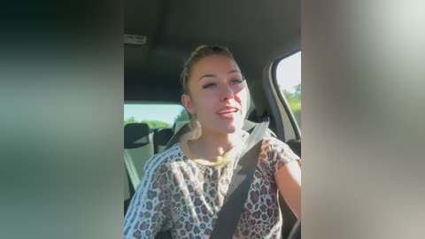 Media: Video of a smiling Caucasian woman with blonde hair, wearing a leopard print top, seated in a car with a seatbelt on, bright sunlight streaming in.