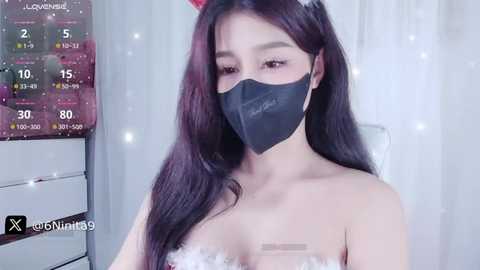 Media: A video of an Asian woman with long black hair, wearing a black face mask, a white strapless dress, and a red headband, standing against a white curtain background with a calendar and a \"XoXoVanity9\" watermark.