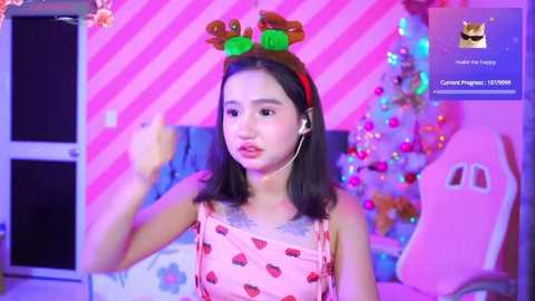 Media: Video of an Asian girl in a pink strawberry-patterned dress, wearing headphones, standing in a pink-striped room with a decorated Christmas tree and a pink gaming chair.