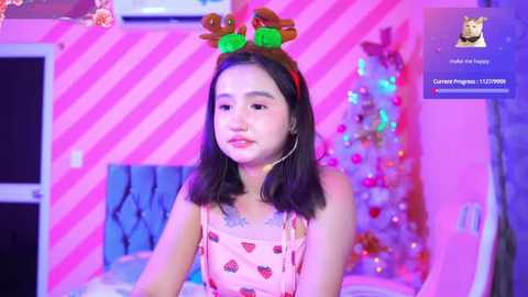 Media: Video of a young Asian woman with shoulder-length black hair, wearing a pink dress with strawberries and a reindeer headband, in a pink-striped room with a decorated Christmas tree.