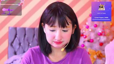 Media: Video of a young Asian woman with straight black hair and fair skin, wearing a purple shirt, listening to headphones, in a pink and orange striped room with a Christmas tree and a bed.