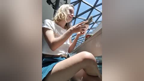 Media: Video of a young, fair-skinned, blonde woman in a white T-shirt and blue skirt, sitting on a chair, reading a book under a sunlit skylight.